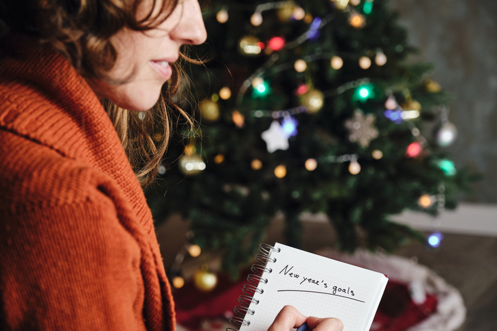 Caucasian woman smiling with orange sweater in profile with new year's goals notebook in hand with unfocused christmas tree in the background