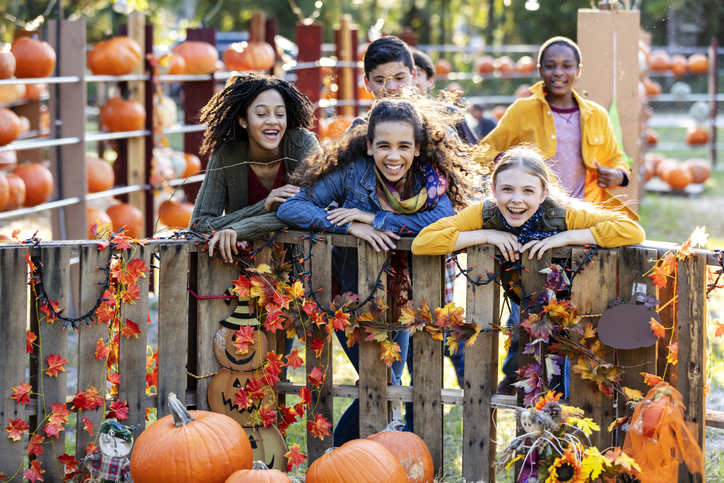 A multi-ethnic group of children having fun at a pumpkin patch. The focus is on the three girls leaning on the wood fence, laughing and looking toward the camera. They are surrounded by pumpkins.